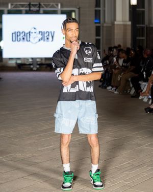 Model wearing light denim shorts and green sneakers poses on runway during fashion show.
