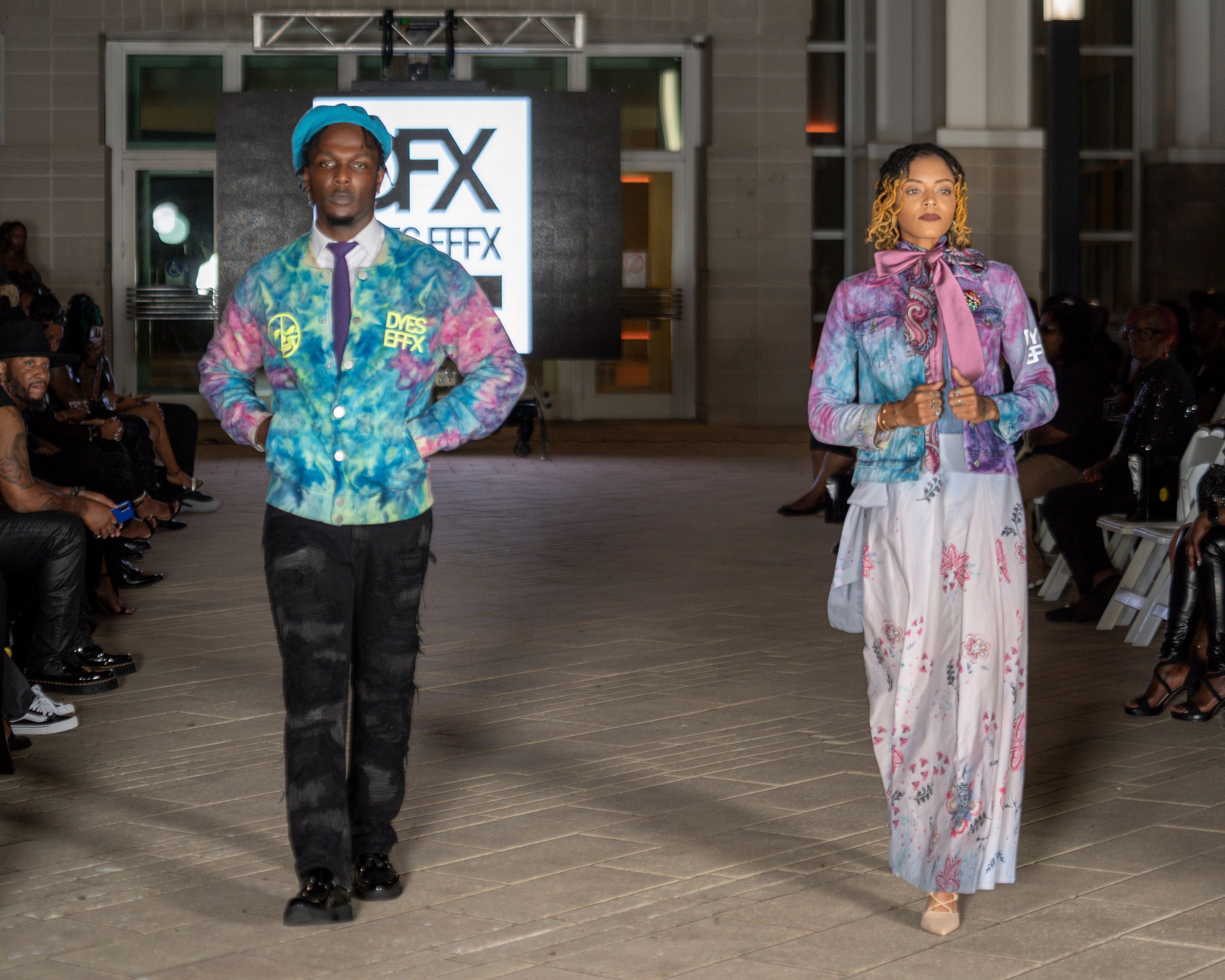 Models showcase colorful pastel prints and dark pants combinations at indoor fashion show.