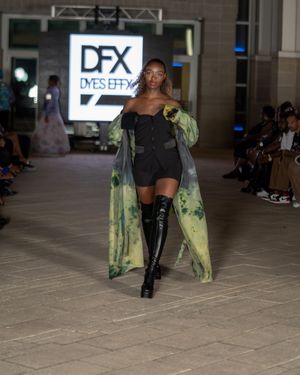 Model wears black mini dress with floral print coat and knee-high boots on runway.