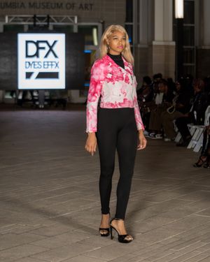 Model showcases pink tie-dye top with black pants during fashion show at museum venue.