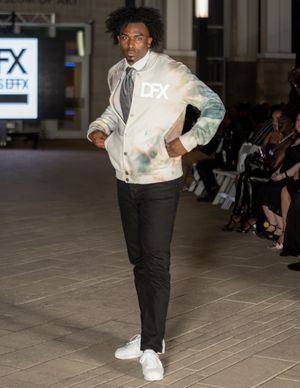 Model showcases white and teal gradient bomber jacket with black pants on fashion runway.