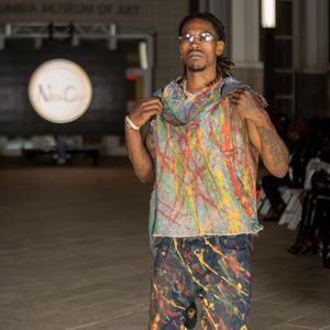 Model showcases colorful paint-splattered outfit with sleeveless top and shorts on fashion runway.