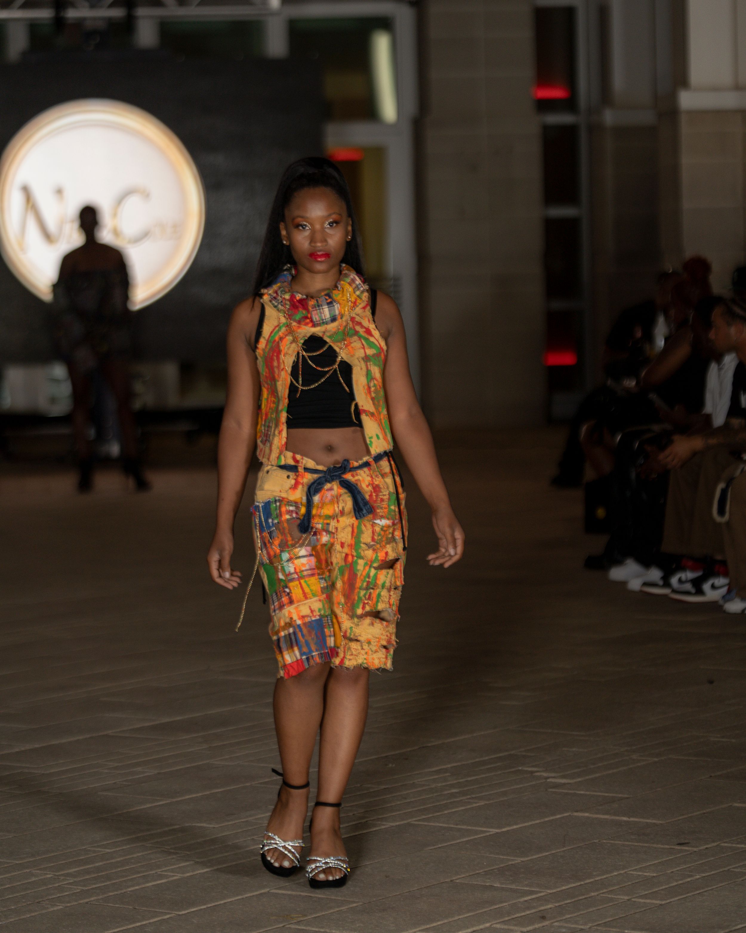 Model walks runway in vibrant orange patterned shorts and black top during fashion show.