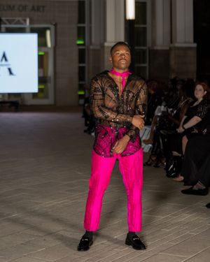 Fashion show featuring hot pink pants paired with sheer black lace top on runway.