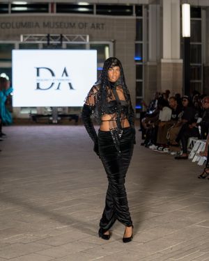 Model showcases black velvet ruched dress on fashion runway at Columbia Museum.