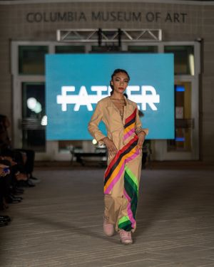 Fashion model walks runway at Columbia Museum of Art wearing colorful striped jumpsuit with beige accents.