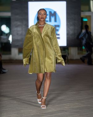 Model walks runway in olive green short jacket and white sandals at museum fashion show.