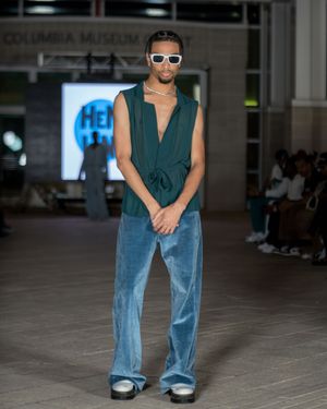 Model in teal sleeveless top and wide-leg jeans walks runway wearing white sunglasses.