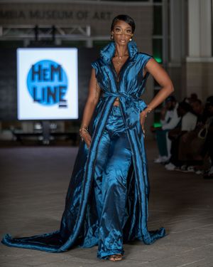Model walks runway in flowing turquoise evening gown with dramatic ruffles at fashion show.