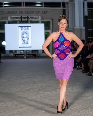 Model walks runway in vibrant purple and blue floral dress at Columbia Museum of Art fashion show.
