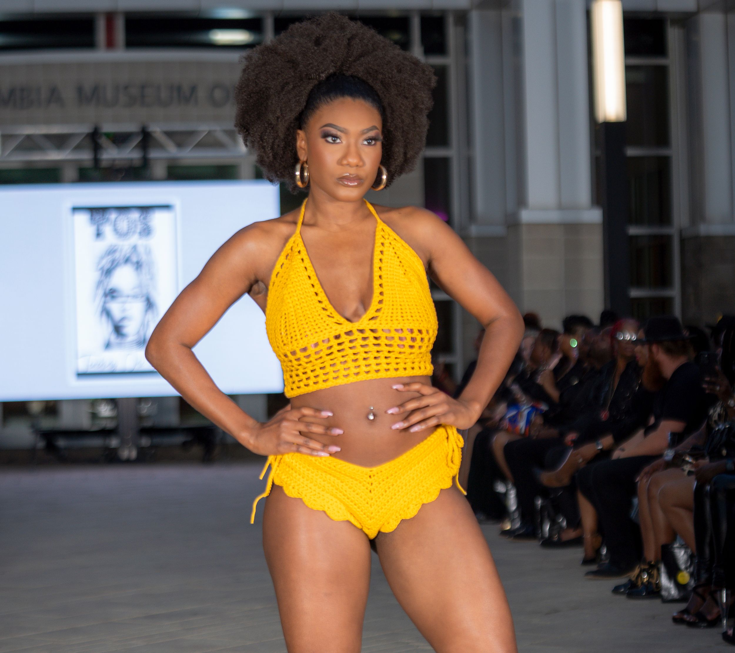 Model walks runway in bright yellow crochet bikini during fashion show at art museum.