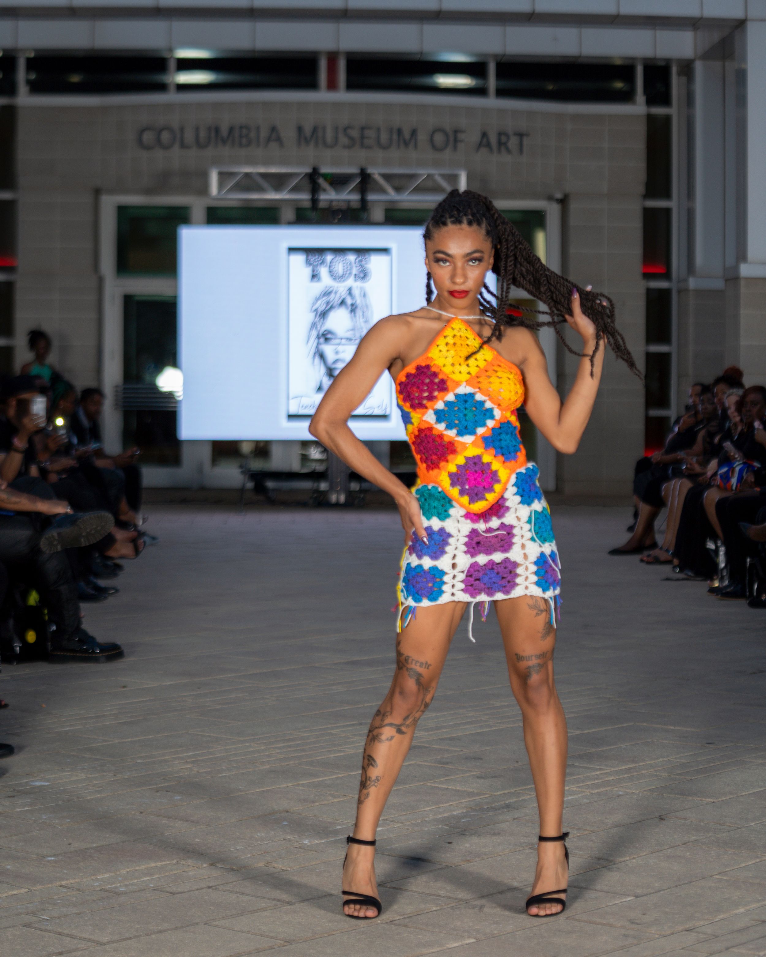 Fashion model wearing colorful floral print outfit walks runway at Columbia Museum of Art fashion show.