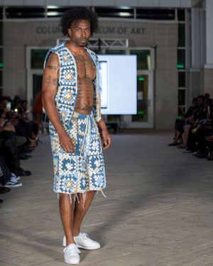 Model walks runway in blue and white patterned vest and shorts ensemble with white sneakers at fashion show.