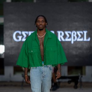 Model walks runway in bright green cropped jacket and light wash jeans with Rebel signage in background at fashion show.