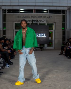 Fashion model showcases green jacket and denim with yellow shoes at Columbia Museum of Art runway show.