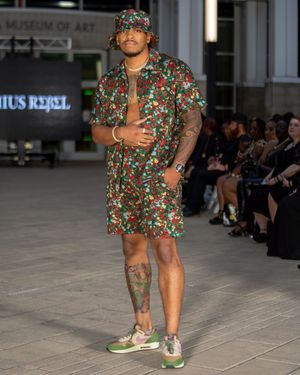 Model walks runway in floral print romper with puff sleeves and green sneakers during fashion show.