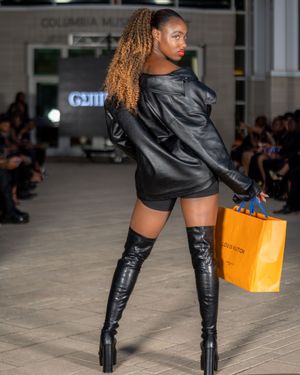 Fashion model walks runway wearing black leather boots and carrying orange shopping bag during fashion show.
