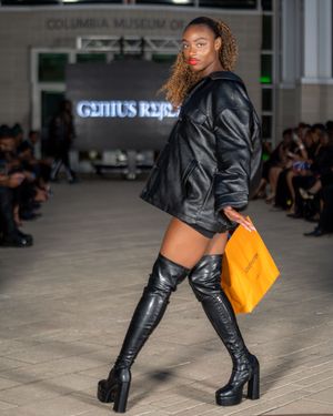 Fashion model walks runway in black leather jacket and boots with bright orange handbag at Columbia Museum of Art show.