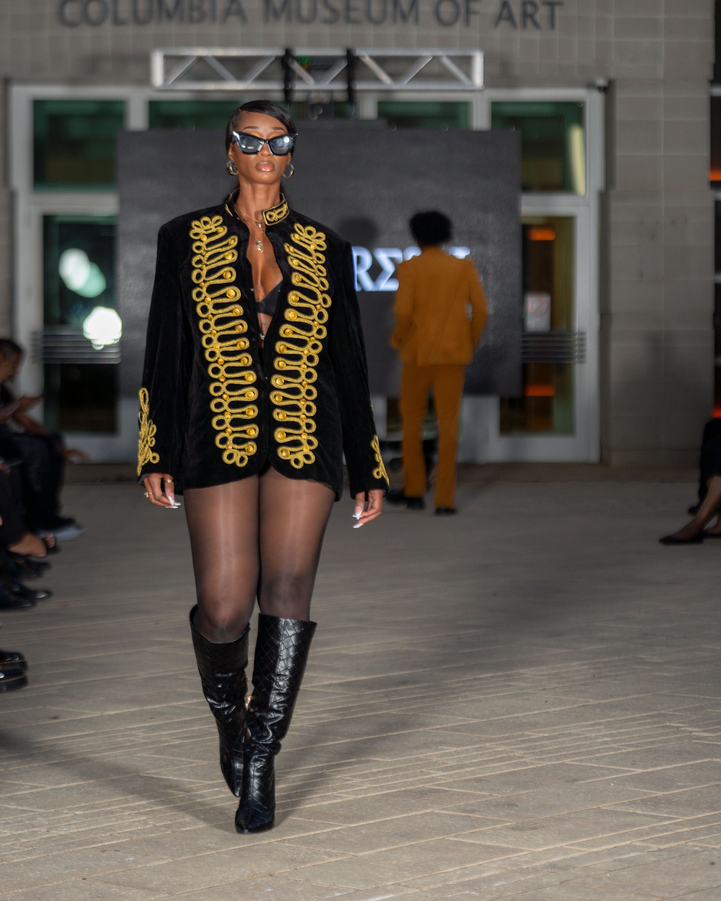 Fashion model struts down catwalk wearing black and gold military-inspired ensemble with knee-high boots.