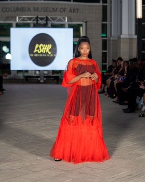 Flowing red sheer gown with dramatic cape effect presented on indoor catwalk.