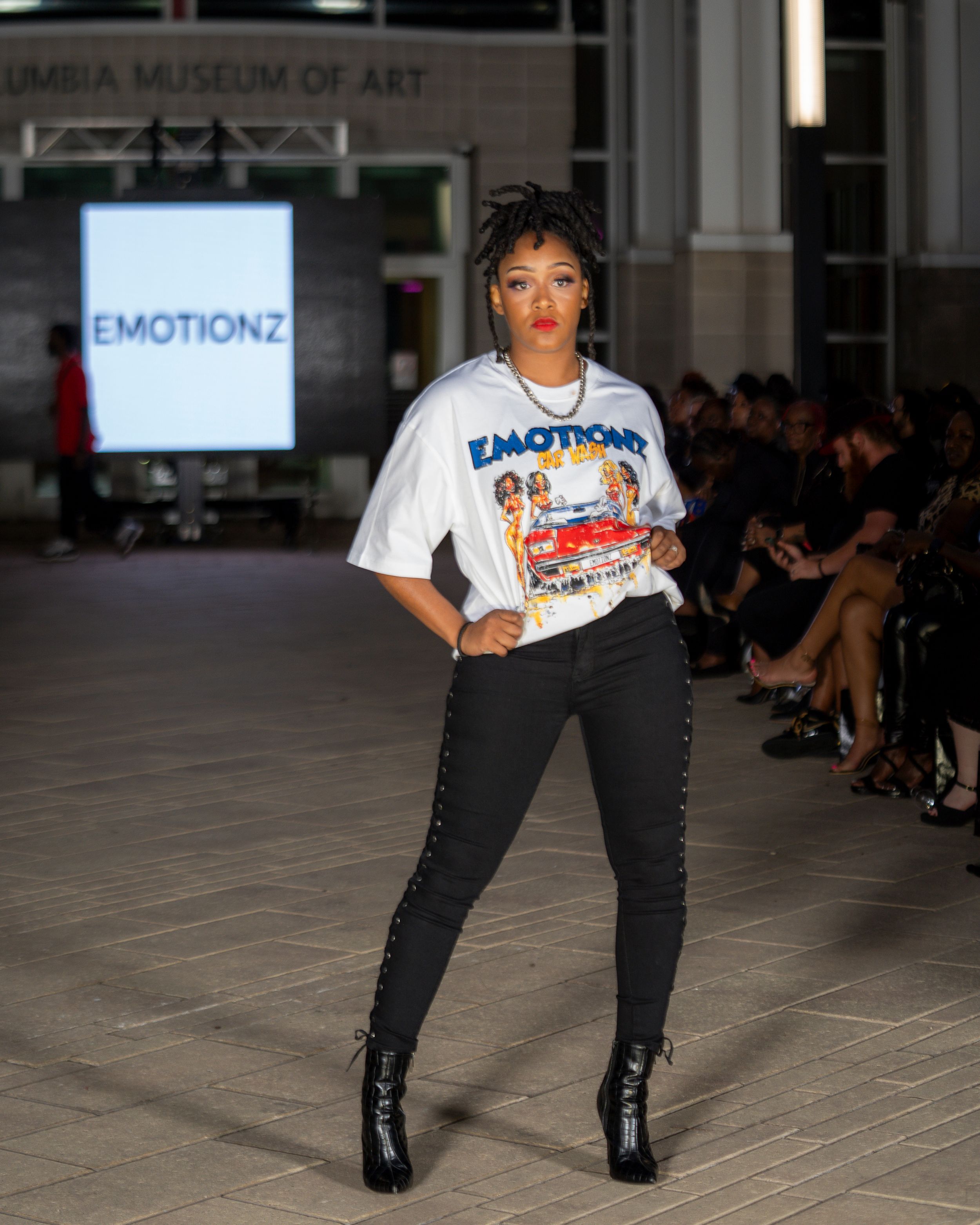 Runway model showcases urban streetwear look with white graphic t-shirt and black pants.