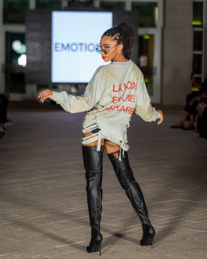Model performs dynamic pose in beige sweatshirt and black leather pants during fashion show.