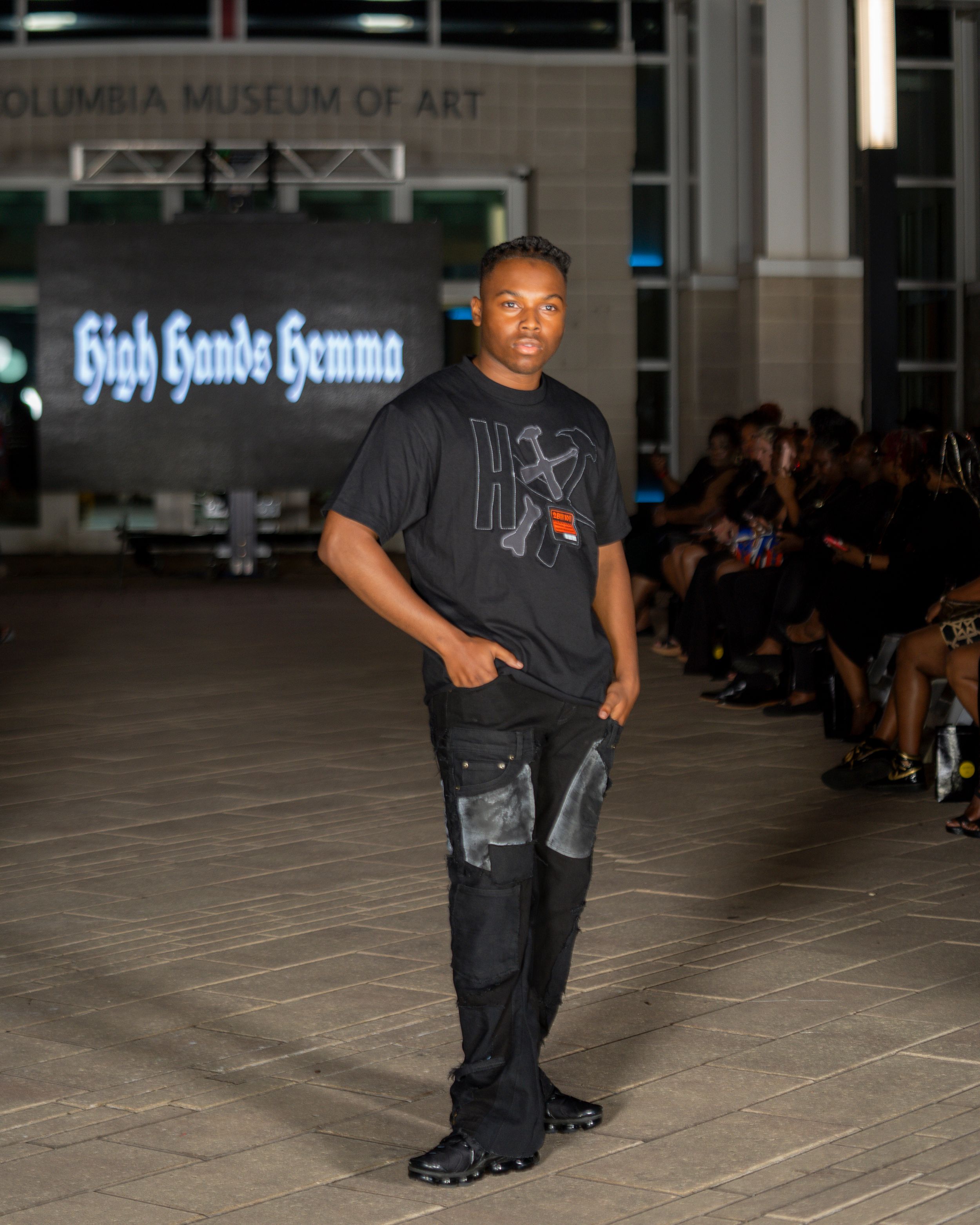 Model presents all-black urban streetwear look with cargo details on concrete runway.