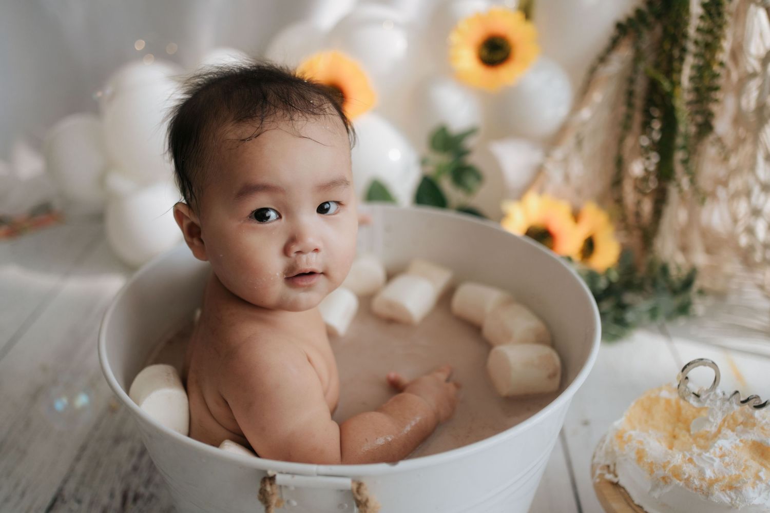 A baby sits in a white chair surrounded by floral decorations and balloons at a birthday celebration.