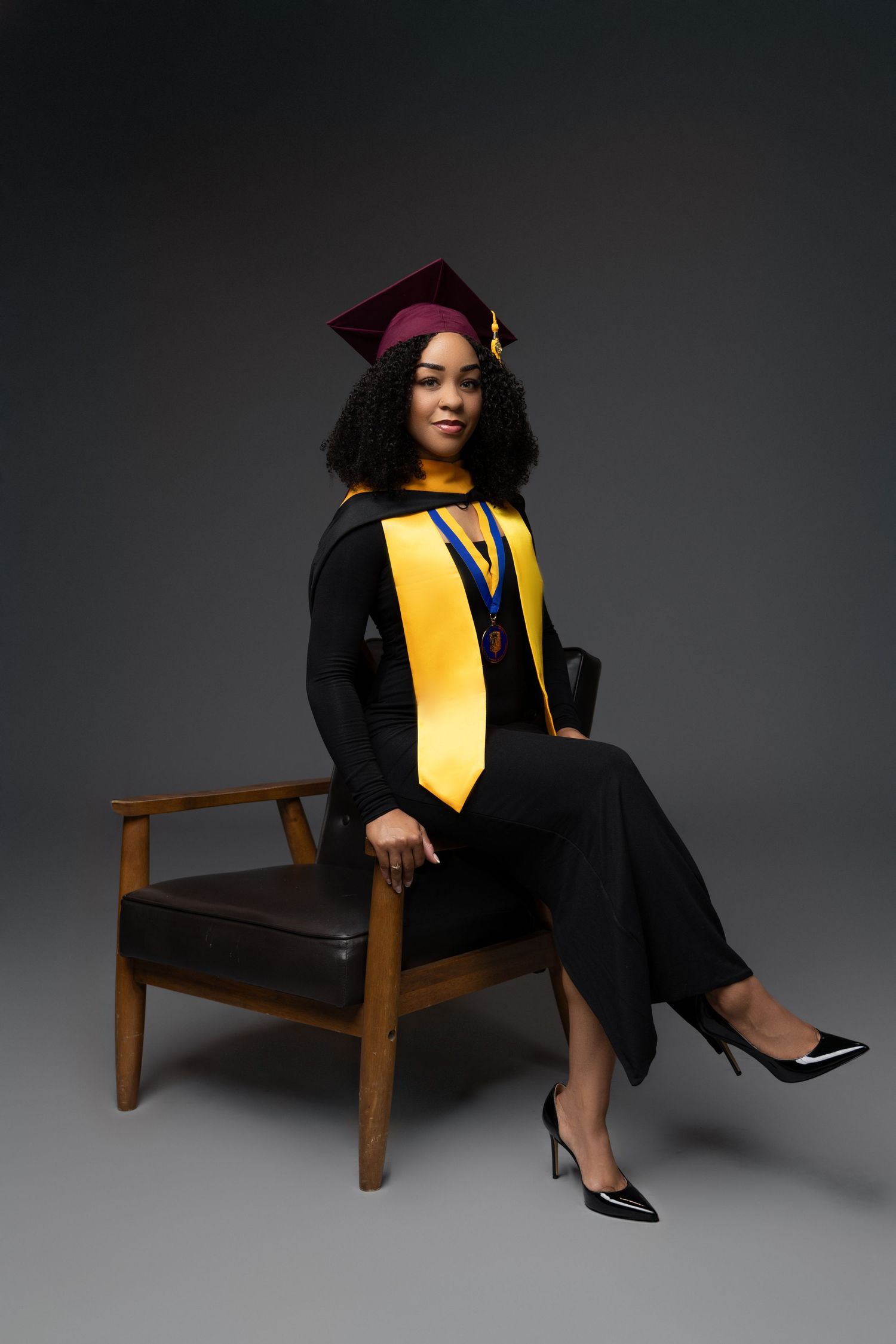 Graduate in black and gold academic regalia poses for professional graduation photos against dark background.