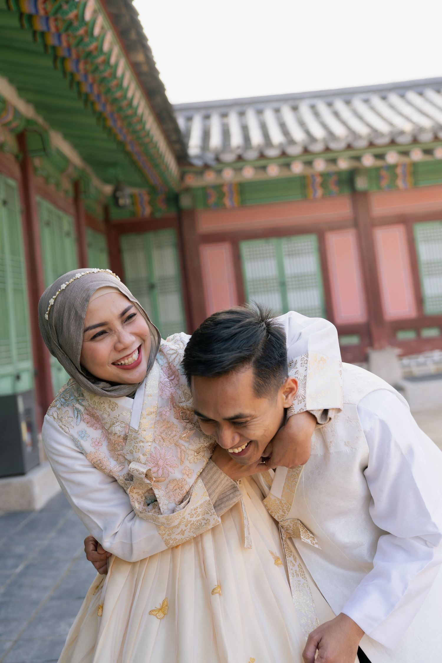 A couple in traditional Korean hanbok attire share intimate moments in front of a palace building with ornate architecture.