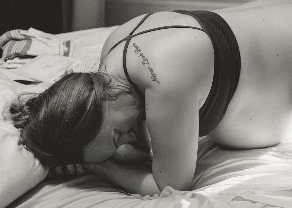 A person in dark clothing lies curled up on white bedding in a black and white photograph.