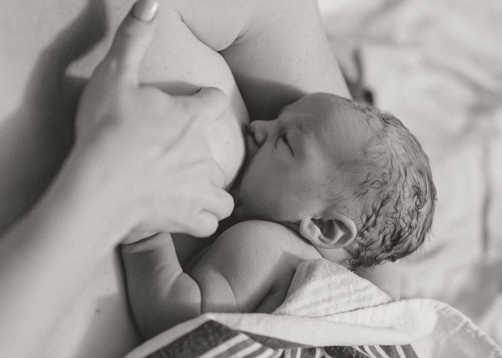 A newborn baby breastfeeds while lying on white bedding in an intimate black and white photograph.