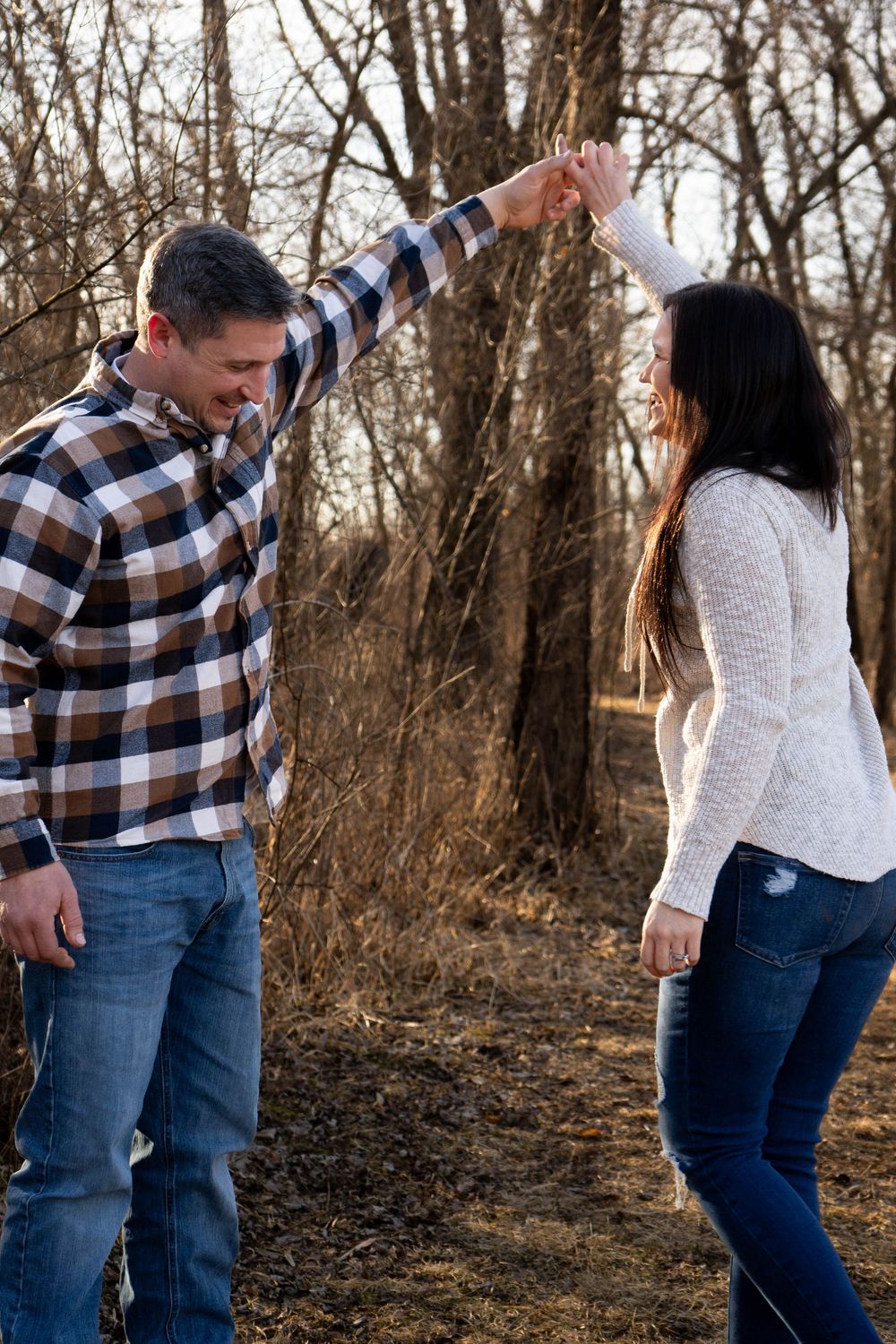 Happy couple dancing and spinning hands together in a wooded area during fall engagement photos.