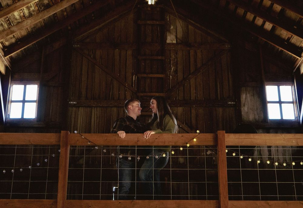 Romantic couple sharing a moment on loft railing inside a barn with string lights.