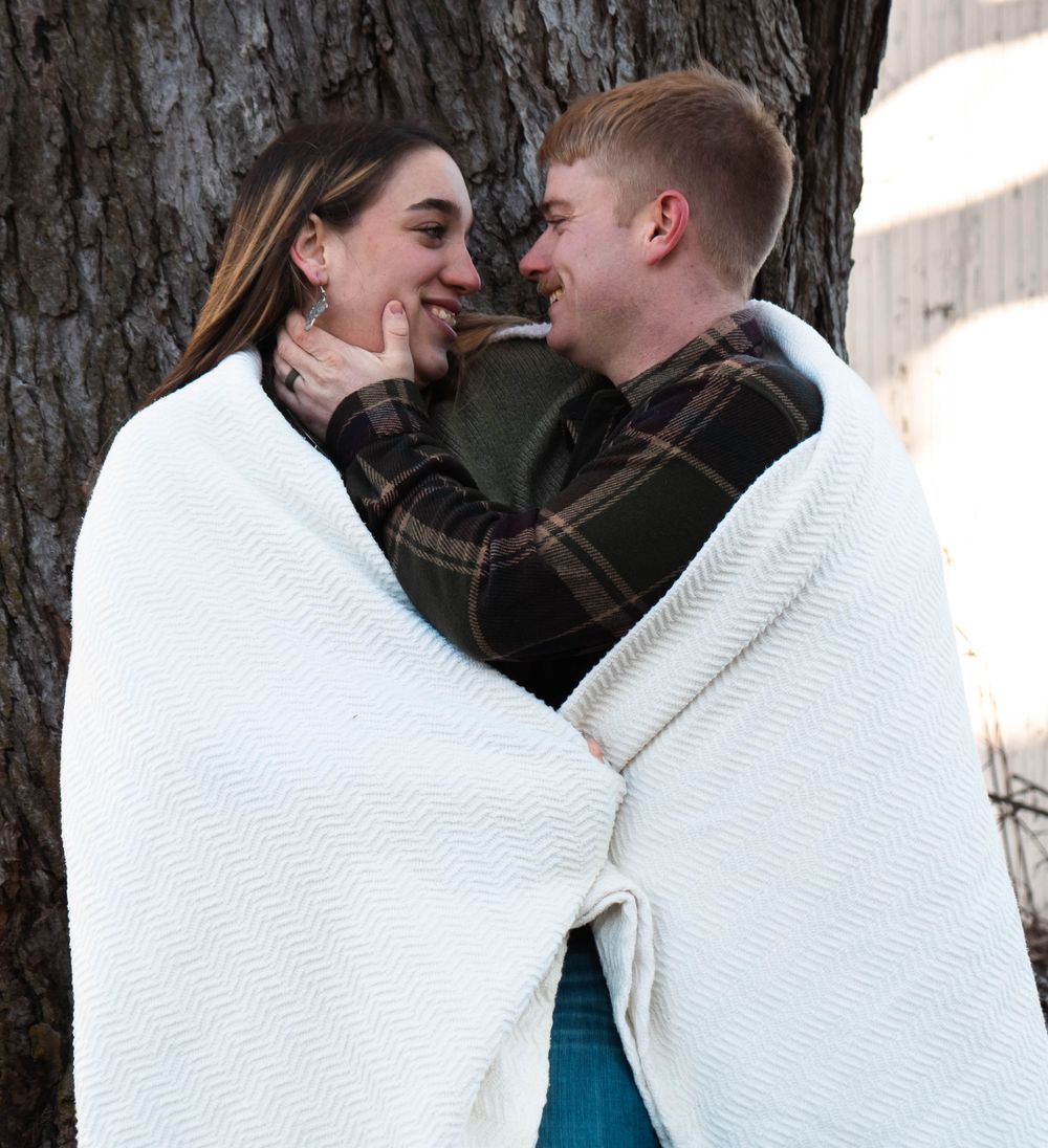 Couple wrapped in white blanket together, smiling and laughing by a large tree.