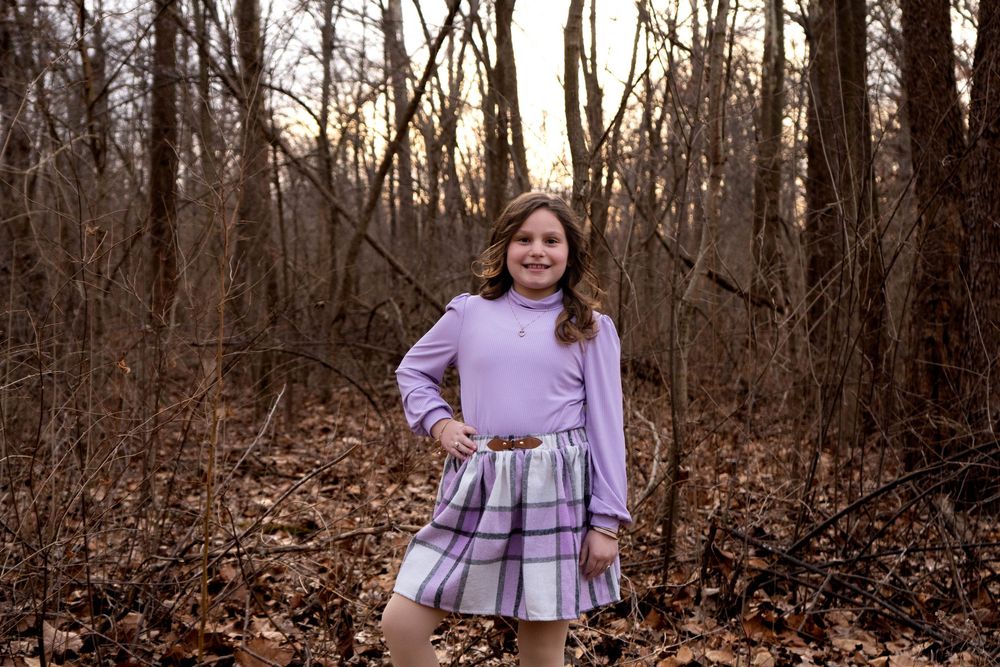 Young girl in purple dress smiling in winter woodland setting with bare trees.