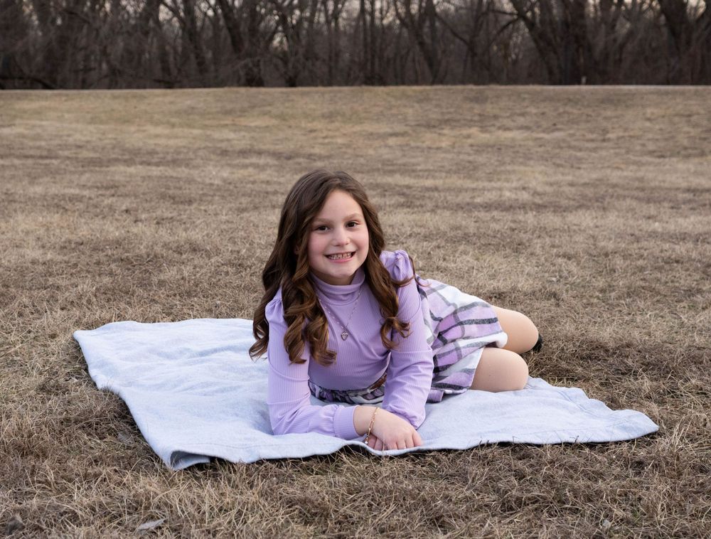 Happy young girl sitting on blanket in open field, wearing lavender outfit.