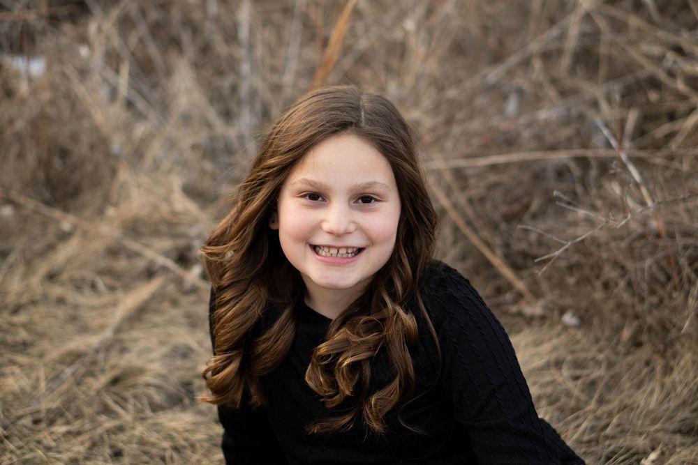 Smiling young girl with curly brown hair posing outdoors among dry brush.