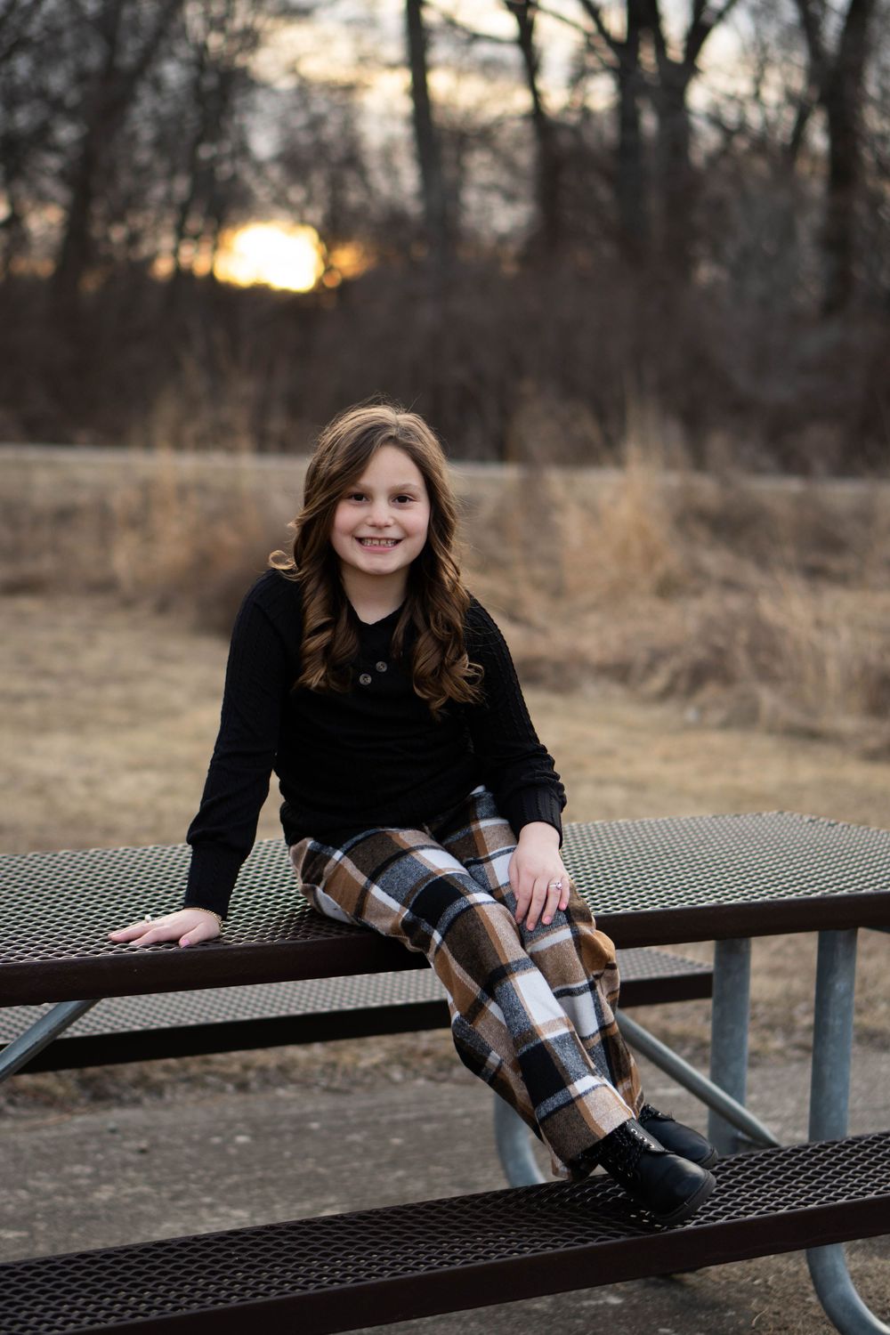 Young girl in black top and plaid pants sitting on picnic table at sunset.