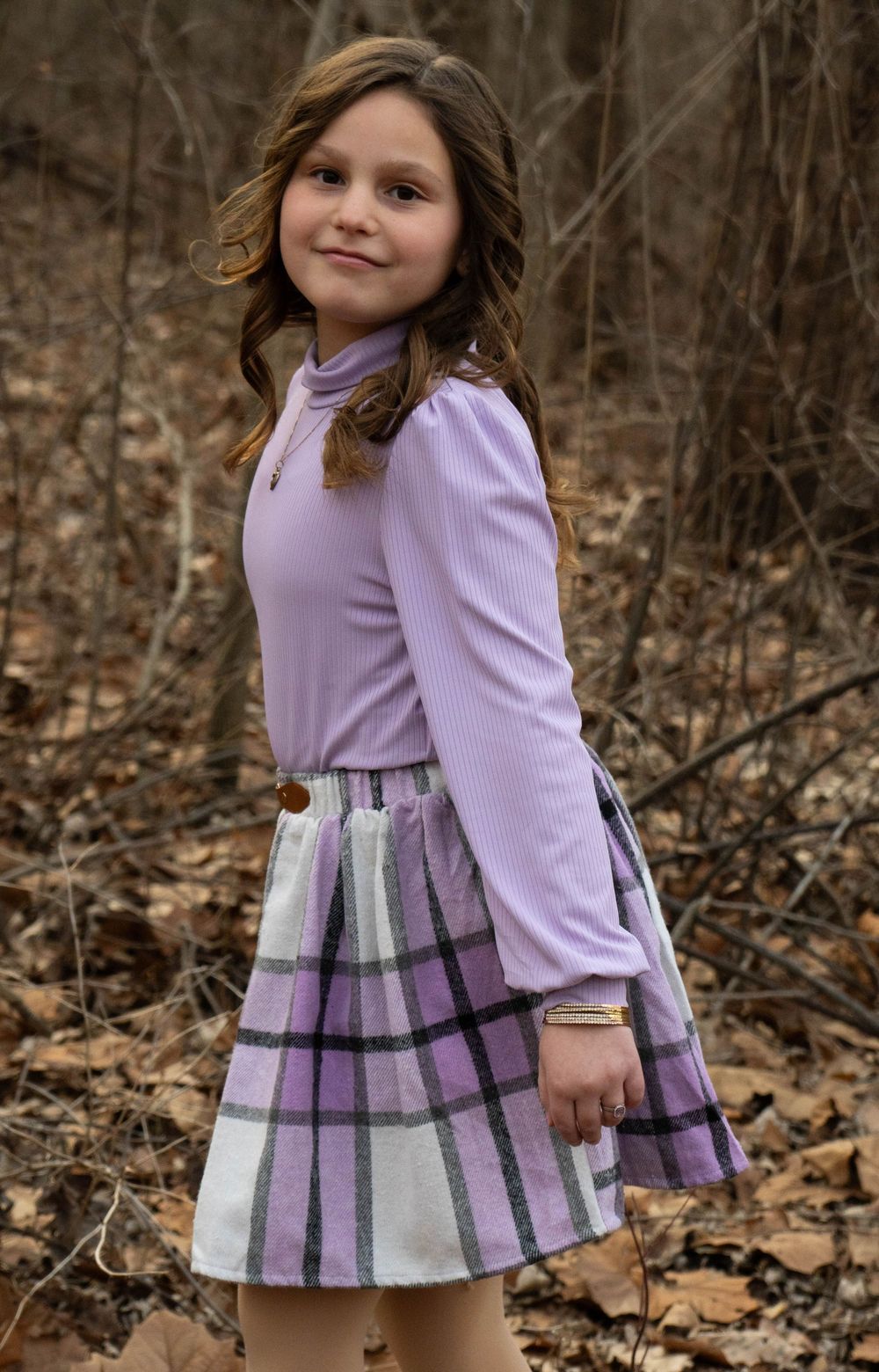 Pretty young girl in purple turtleneck and plaid skirt posing in autumn forest.