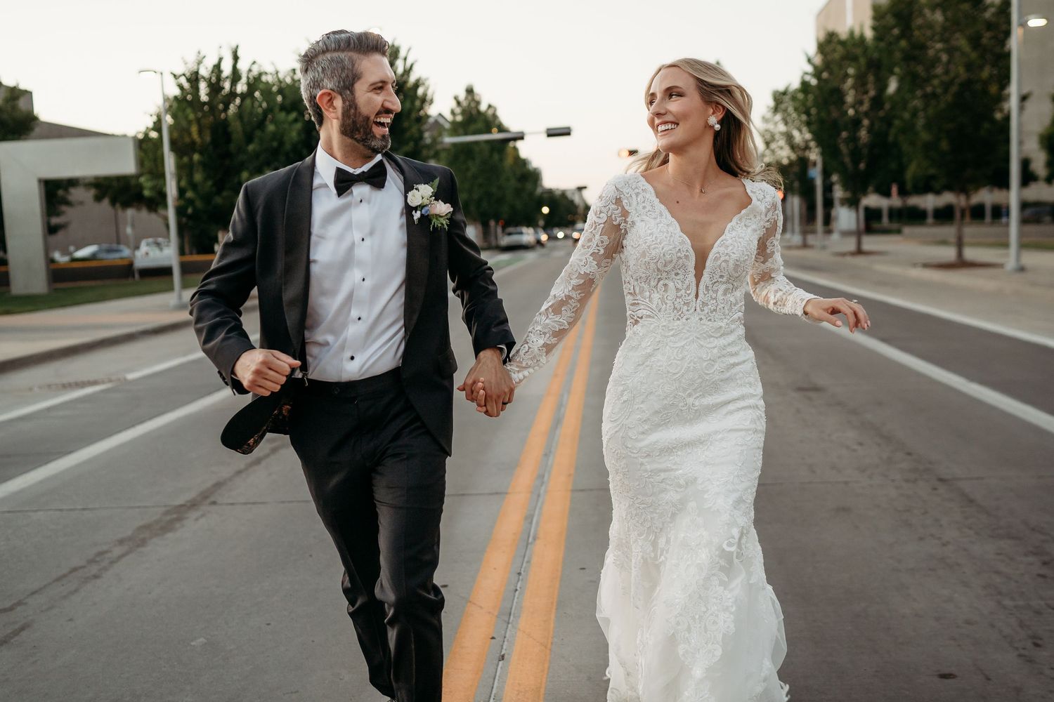 A couple in formal wedding attire runs playfully down an empty street while holding hands.