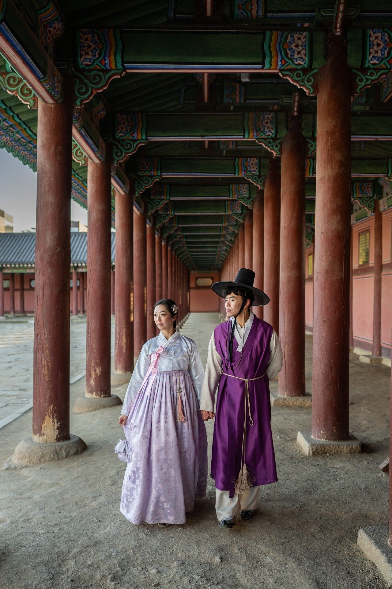 Two people wearing traditional Korean hanbok clothing walk through a covered walkway with ornate red pillars at Gyeongbokgung Palace.