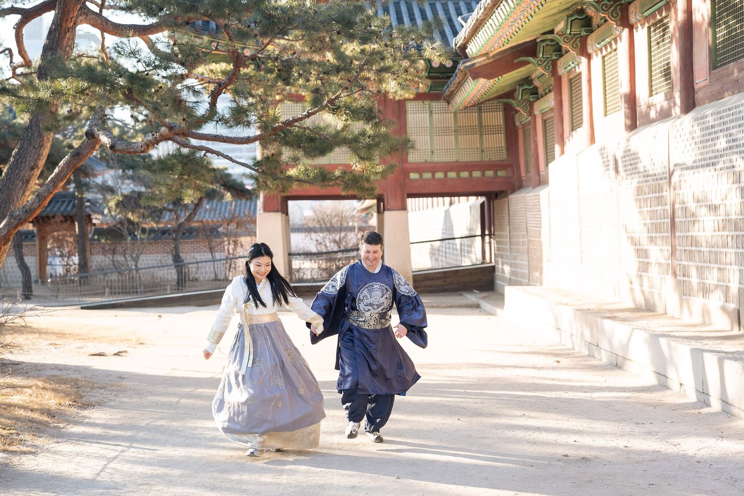 Two people in traditional Korean hanbok clothing run playfully along a temple pathway on a sunny day.