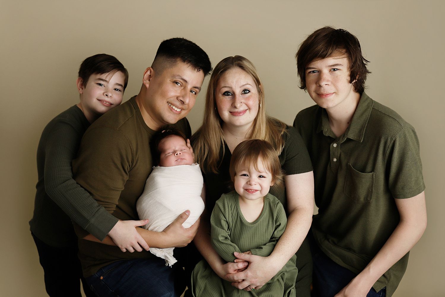 A family wearing coordinating olive green and brown outfits poses together on a neutral backdrop for portraits. Utah County baby photos