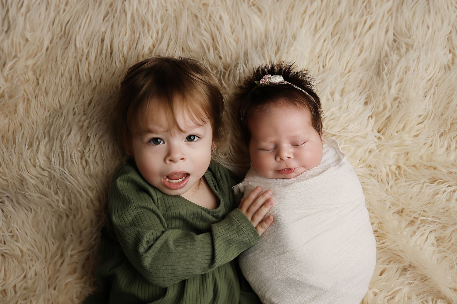 Two siblings pose together on a cream-colored fur rug, with one wearing a green sweater and one swaddled in white. newborn photography