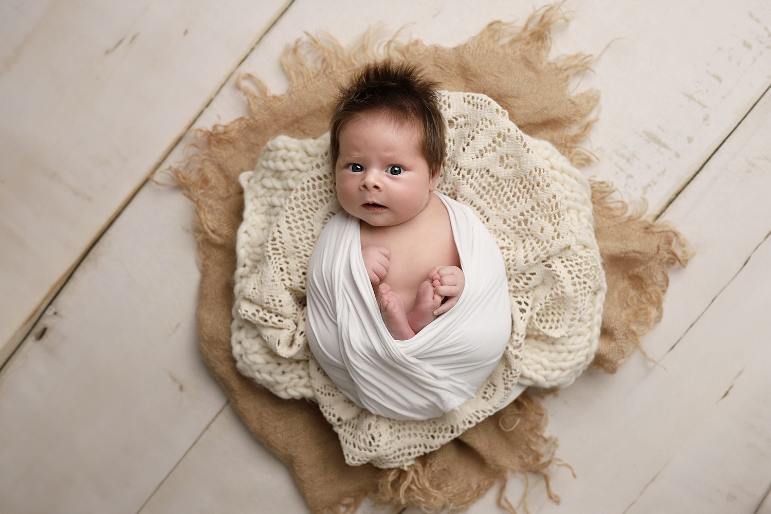 A newborn baby wrapped in white fabric lies on a beige crocheted blanket against a light wooden floor. Newborn photos utah