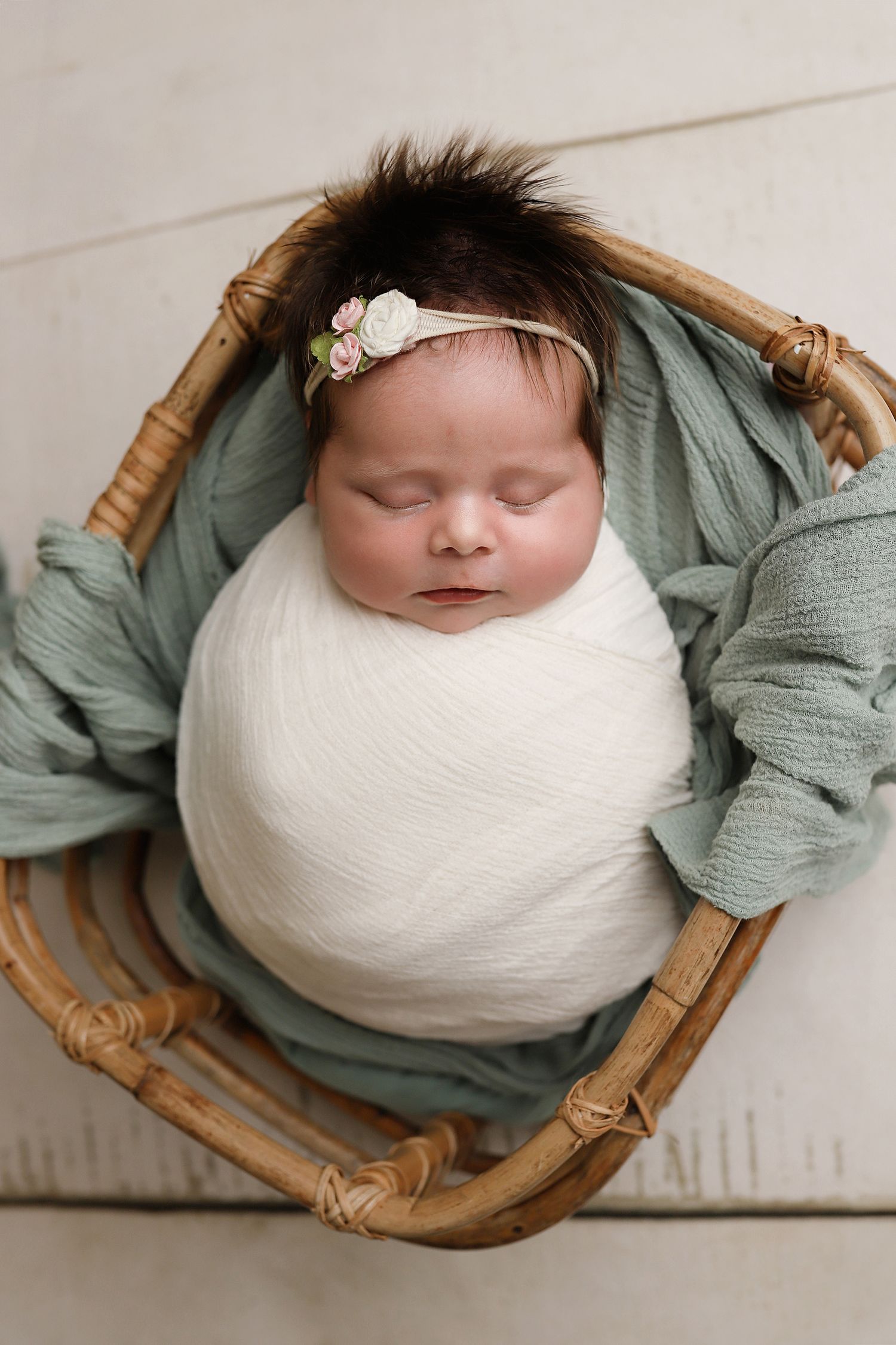 Sleeping newborn wrapped in white swaddle lying in bamboo basket with sage green fabric and delicate floral headband. Orem utah baby photos