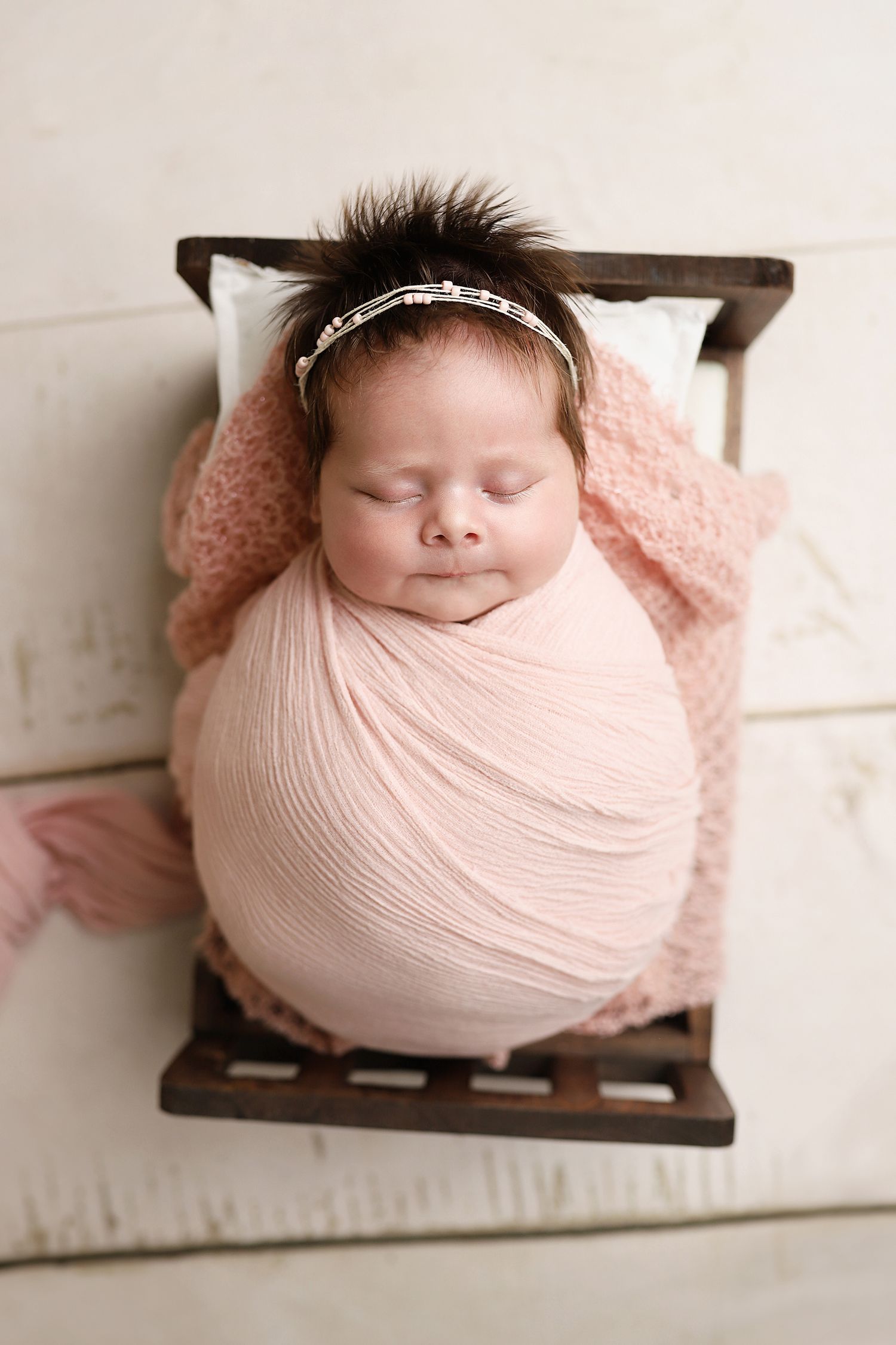 A sleeping newborn swaddled in soft pink fabric lies peacefully on a small wooden bed prop. Utah County newborn photographer