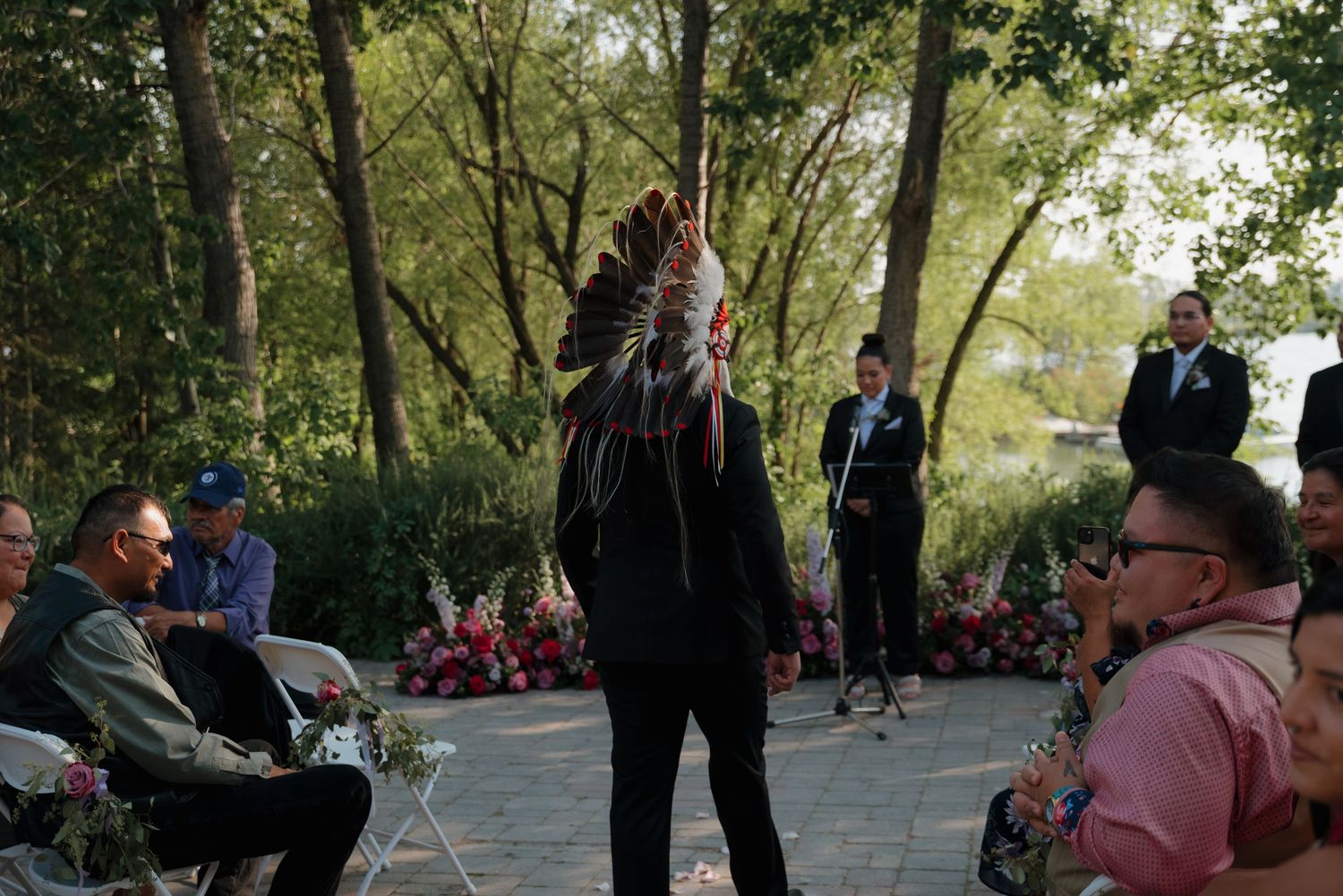 A wedding ceremony takes place in a lush garden setting with guests gathered under trees and greenery.