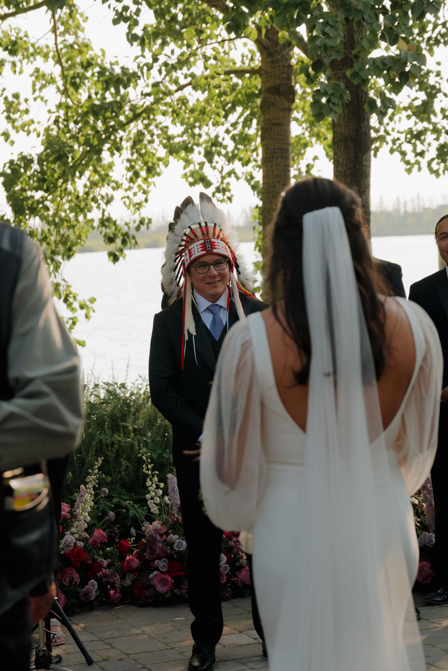 An outdoor wedding ceremony by a lake with a bride in a white dress and traditional Native American headdress.